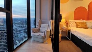 A hotel room with a large window offering a city view, a bed with white bedding, and a chair with a blanket.