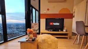 A modern living room with a large window, a flat-screen TV, a wooden coffee table, and a beige pouf.