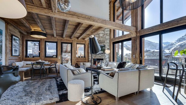 Un salon spacieux avec des poutres en bois, de grandes fenêtres, une cheminée et une vue sur des montagnes enneigées.