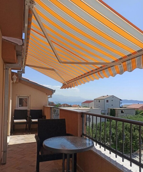 A balcony with a striped awning, a round table, two chairs, and a view of the sea and buildings in the background.