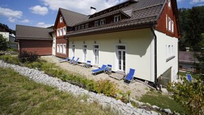 A two-story building with a sloped roof, a covered outdoor seating area with blue chairs, and a stone retaining wall. - Přehrada Souš by Interhome (Desná v Jizerských horách)