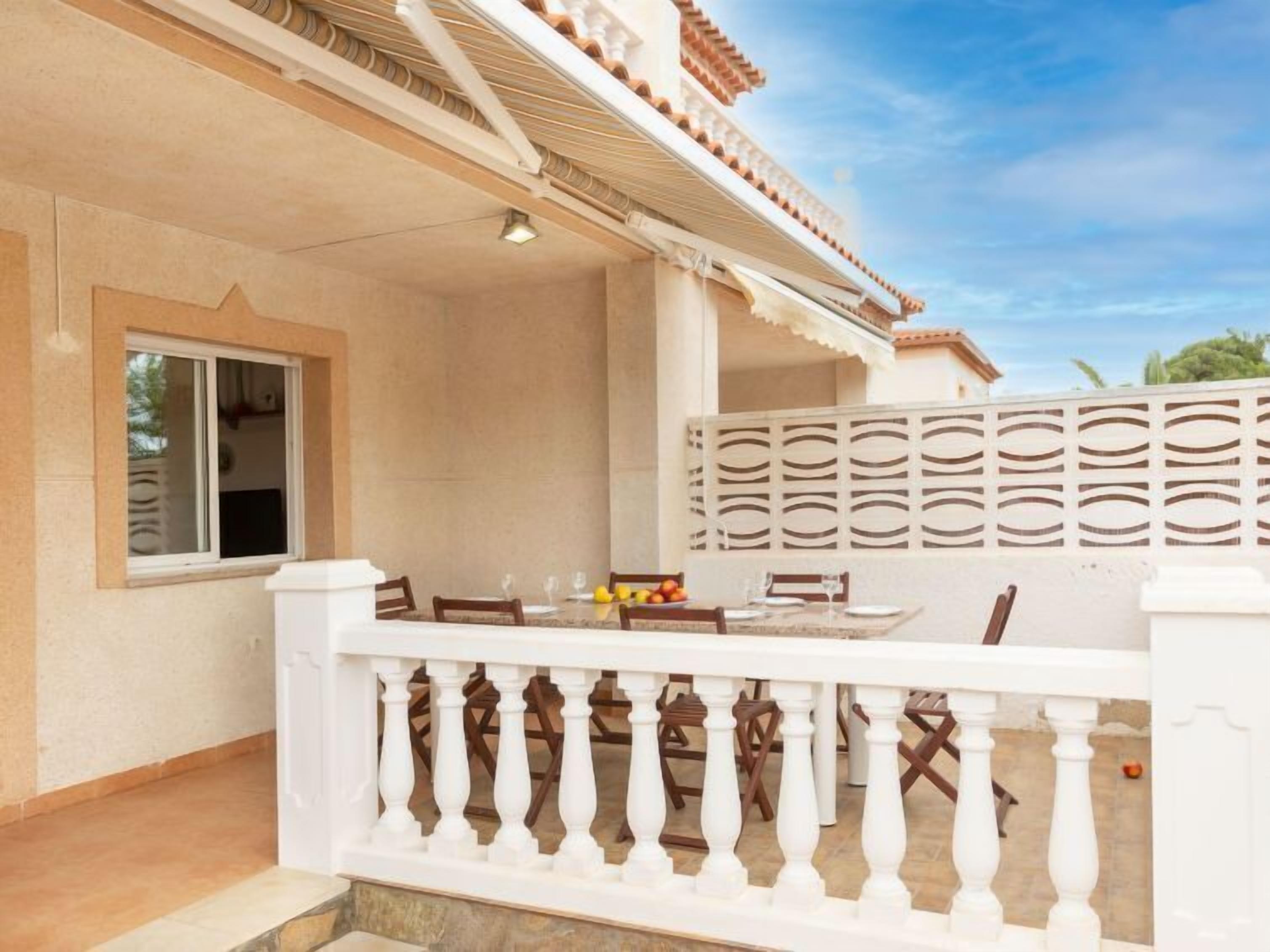 A covered outdoor dining area with a table set for two, a white balustrade, and a view of the sky.
