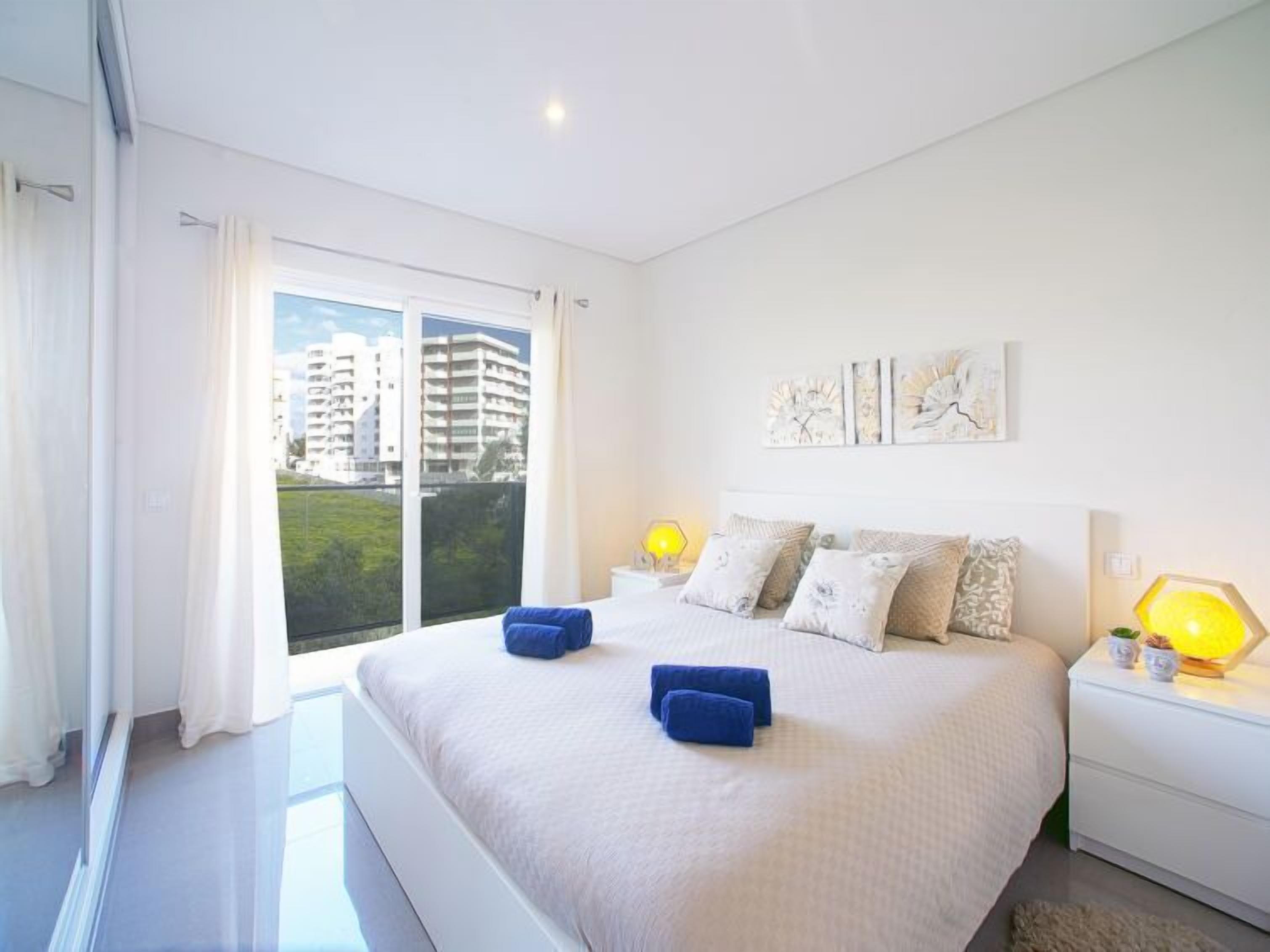 A modern bedroom with a large bed, two bedside lamps, and a view of buildings through the window.