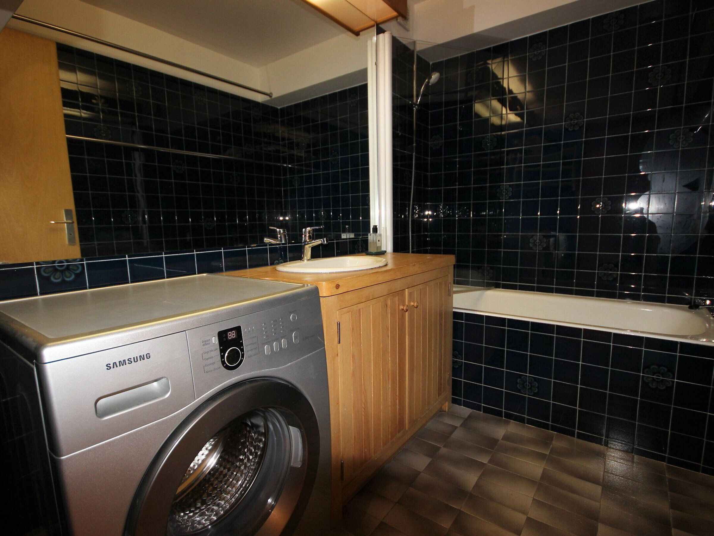 A compact laundry area with a Samsung washing machine, a wooden cabinet, a sink, and a bathtub.