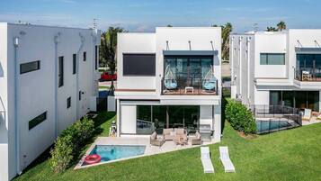 Una casa moderna de dos pisos con piscina, mobiliario exterior y un balcón con sillones colgantes.