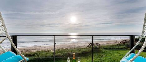A balcony with wicker chairs and a table, overlooking a beach with a wooden deck and a fence.