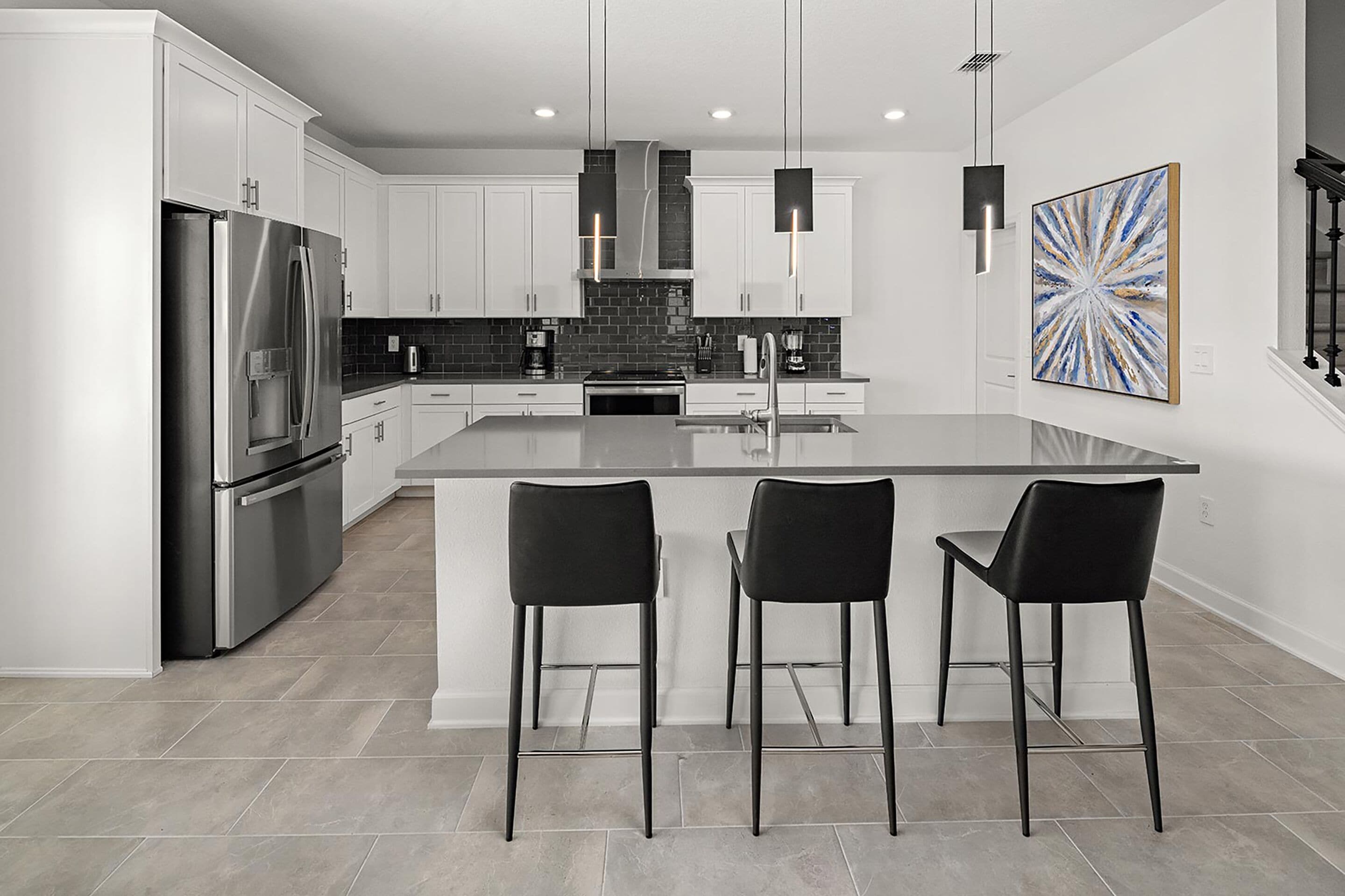 A modern kitchen with white cabinets, a stainless steel refrigerator, a large island with bar stools, and pendant lighting.