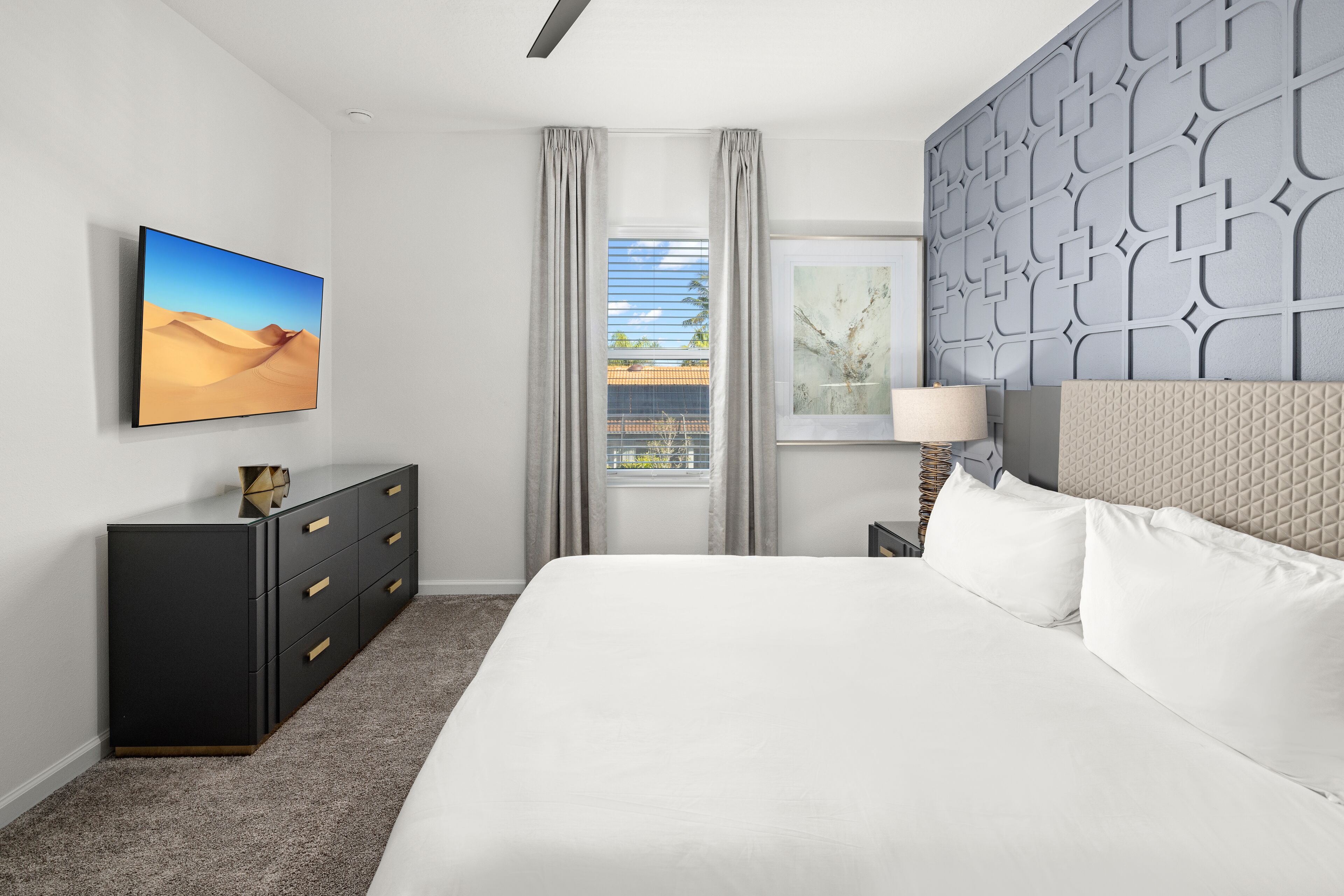 Ein modernes Hotelzimmer mit einem großen Bett, einer Kommode, einem Gemälde von einer Wüstenlandschaft, einem Fenster mit Vorhängen und einer strukturierten Wandverkleidung.