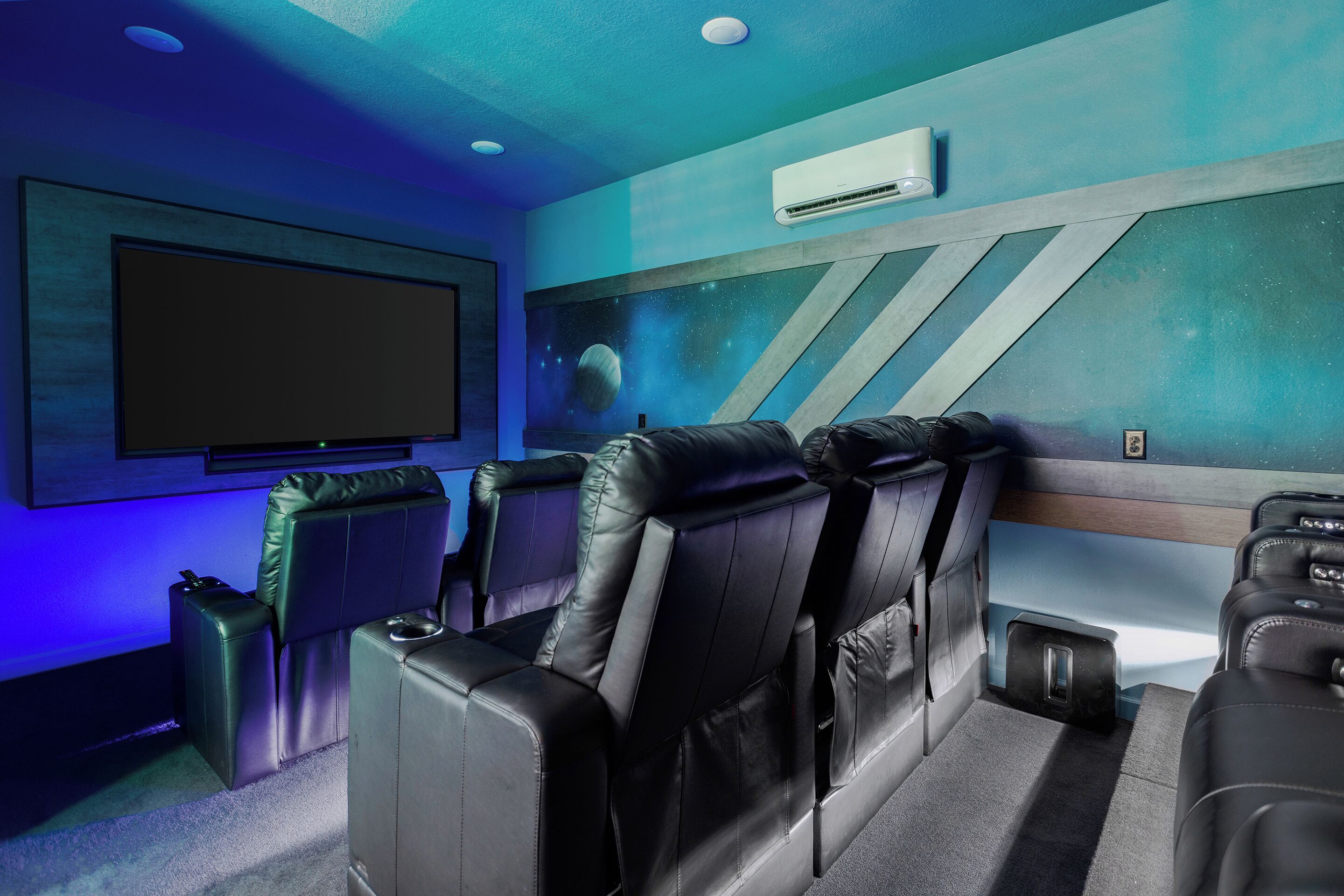 Sala de cine en casa con butacas reclinables de cuero negro, un televisor de pantalla plana grande y un mural espacial en la pared.