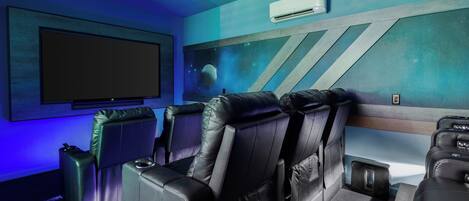 Sala de cine en casa con butacas reclinables de cuero negro, un televisor de pantalla plana grande y un mural espacial en la pared.