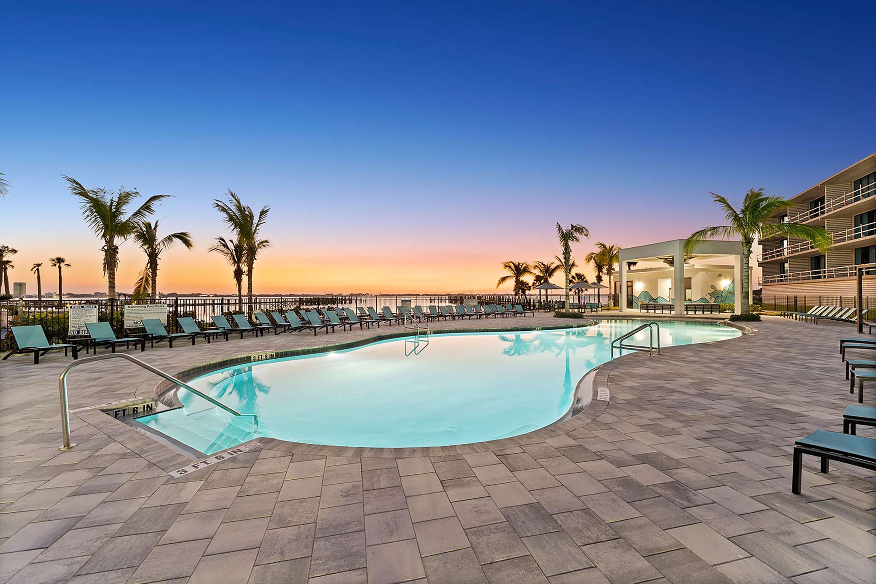 A pool area with lounge chairs and palm trees, surrounded by a building with multiple floors.