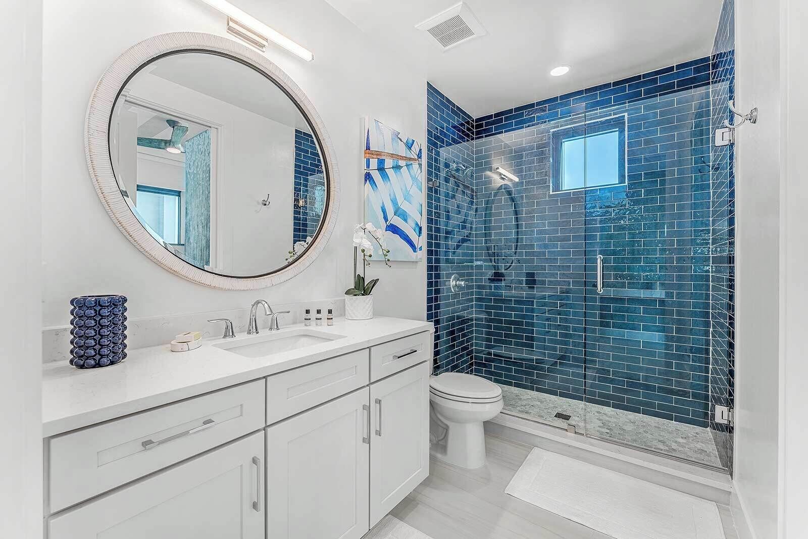 A modern bathroom with a white vanity, a round mirror, and a glass shower enclosure with blue mosaic tiles.
