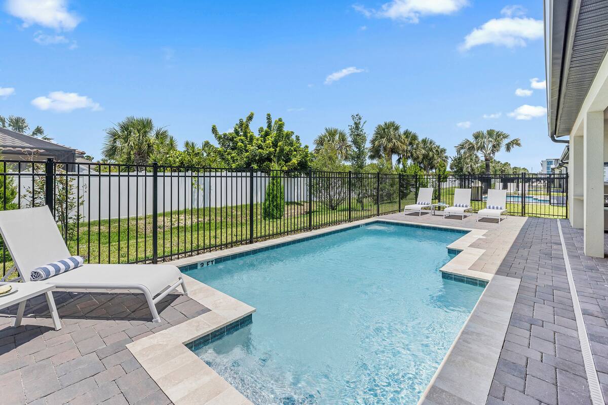 Ein rechteckiger Swimmingpool mit schwarzem Metallzaun, umgeben von einer gepflasterten Terrasse mit Liegestühlen und einer Rasenfläche mit Palmen.