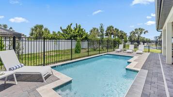 A rectangular swimming pool with a black metal fence, surrounded by a paved patio with lounge chairs and a grassy area with palm trees.