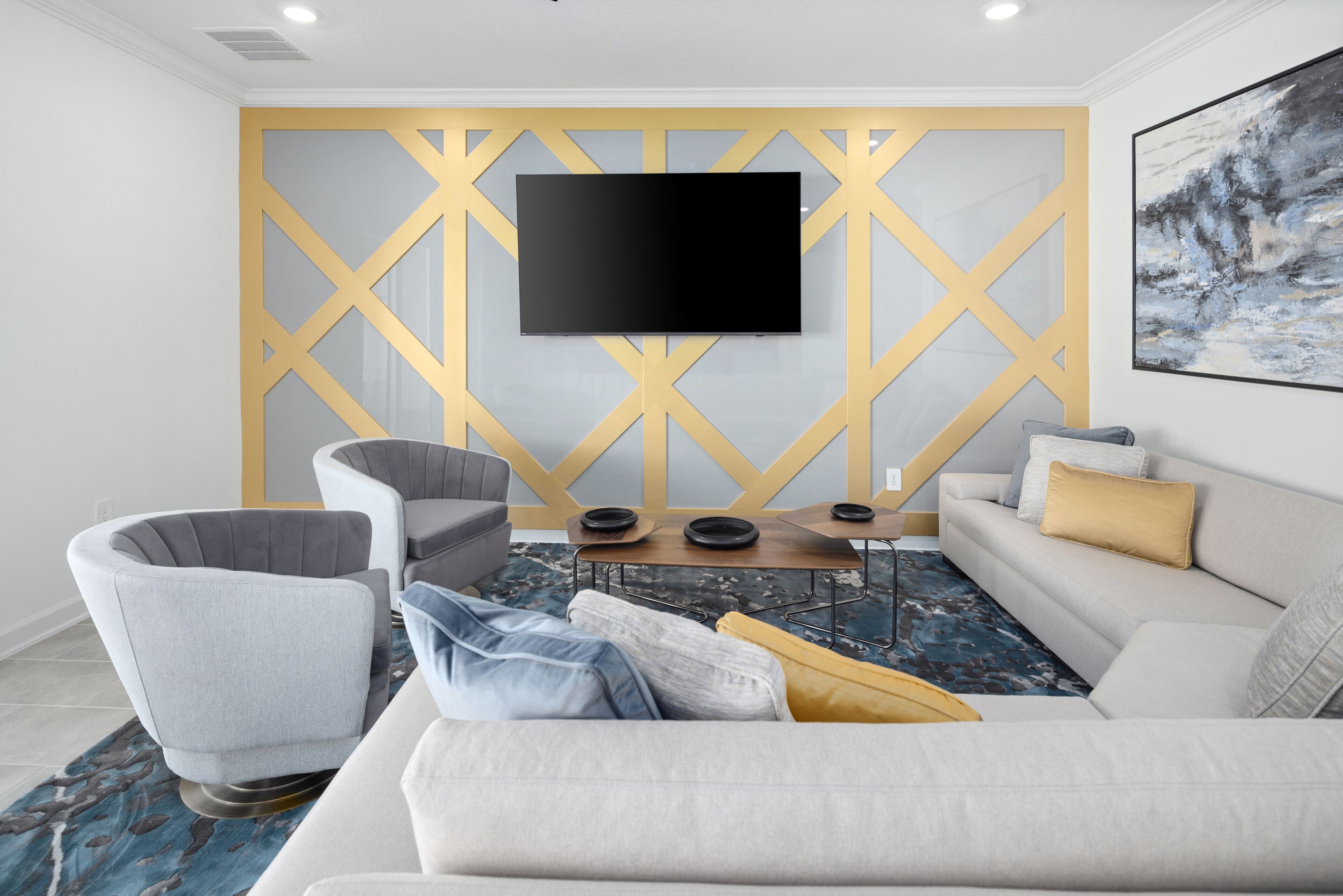 A modern living room with a flat-screen TV mounted on a geometric wall, a grey armchair, a sofa with cushions, and a coffee table with a blue patterned rug.