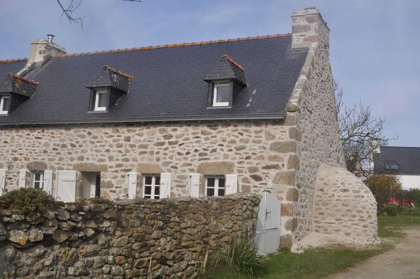 Een stenen huis met schuin dak, meerdere ramen en een stenen muur ervoor.