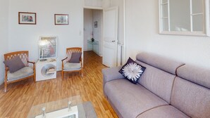 Ein Wohnzimmer mit einer Couch, zwei Sesseln, einem Couchtisch und einem kleinen Beistelltisch. An der Wand hängen gerahmte Bilder, eine Tür führt in einen anderen Raum, und ein Fenster mit weißem Rahmen ist zu sehen.