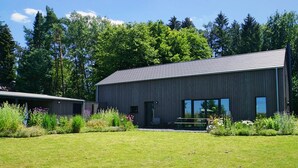 A modern building with a sloped roof and large windows, surrounded by a well-maintained lawn and various plants. - Wäller Haus - Design home with fireplace & private fenced garden (Breitscheid)