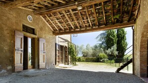 Interior abierto y rústico, con suelo de grava, vigas de madera y vistas a un jardín con árboles y una valla.