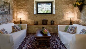 Una habitación rústica con paredes de piedra, dos sofás blancos con cojines florales, una mesa de madera con un jarrón de flores y una ventana que ofrece vistas a la vegetación.