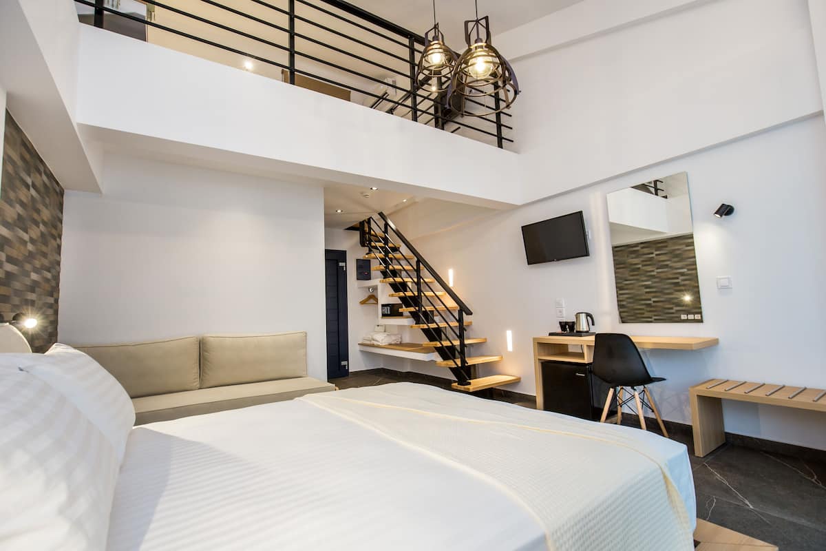 A modern hotel room with a large bed, a desk, a chair, a small table, a TV, a mirror, and a staircase leading to an upper level.