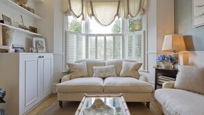 A living room with a beige sofa, a glass coffee table, and a window with shutters. - Cayenne Canopy, by Veeve (London)