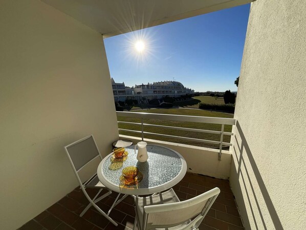 A small balcony with a table and chairs, a white pitcher, and a view of a hotel and golf course.