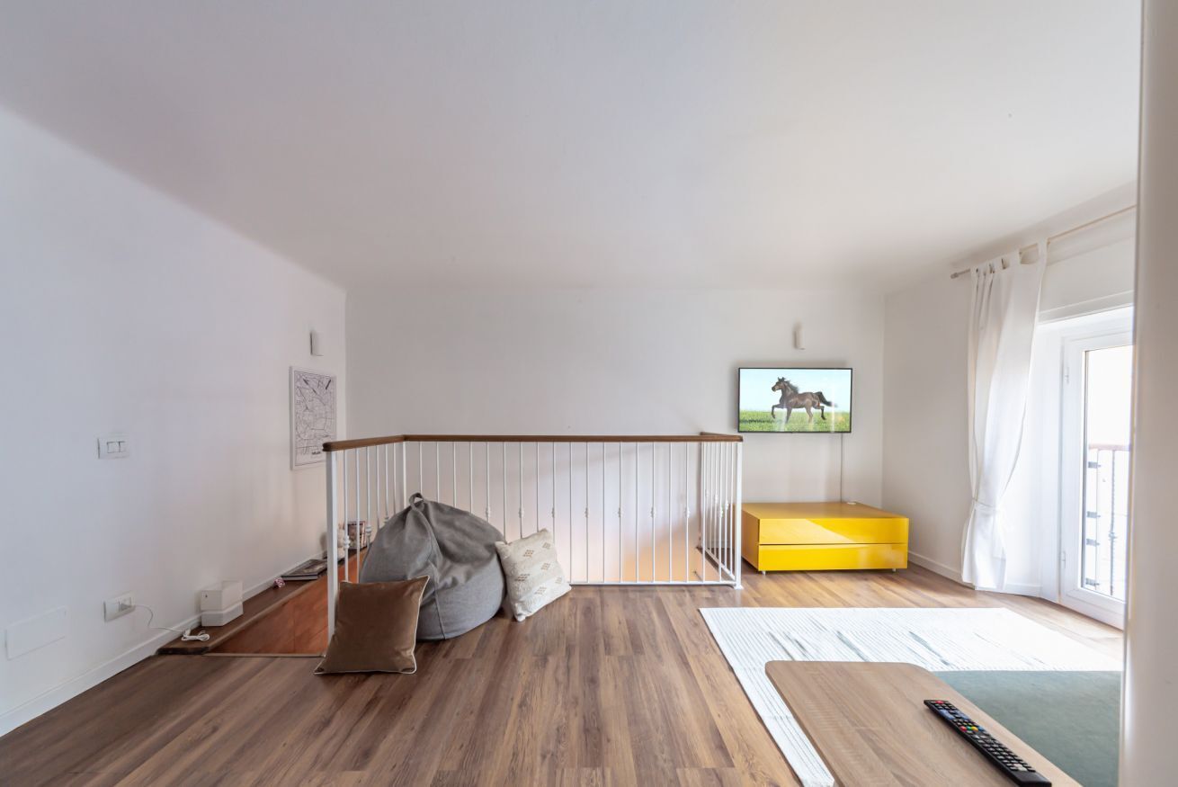 Una sala de estar moderna con piso de madera, un sofá amarillo, un asiento informal gris, una mesa de centro de madera, un televisor montado en la pared y una ventana con cortinas.