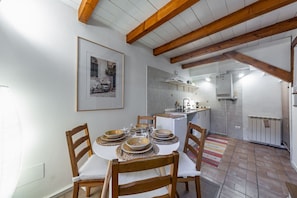 A dining area with a round table set for four, wooden chairs, a kitchen in the background, and exposed wooden beams on the ceiling.