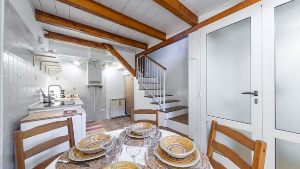 A dining area with a table set for four, wooden chairs, a staircase, and a kitchen in the background.