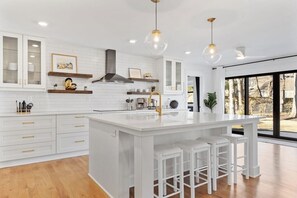 Una cucina moderna con mobili bianchi, una grande isola con sgabelli da bar, illuminazione a sospensione e vista attraverso ampie finestre.