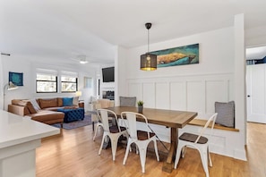 A modern living room with a wooden dining table, white chairs, a brown leather sofa, and a hanging light fixture.