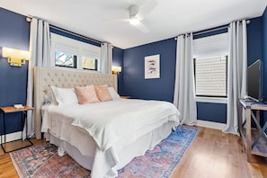 A bedroom with a tufted headboard bed, white bedding, a patterned rug, a ceiling fan, and a television on a wooden stand.