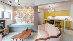 A modern living room with a sofa, a coffee table, and a TV. There is a kitchen area with yellow cabinets and a bar counter. - Acropolis Pop Art Apartment (Athens)