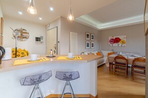 A modern kitchen with a central island, bar stools, a dining area with a table and chairs, and a living area with a sofa.