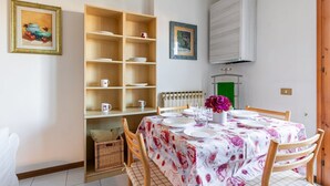 Uma área de jantar com uma mesa posta para quatro pessoas, cercada por cadeiras de madeira. Há uma estante com diversos objetos e duas fotos emolduradas na parede.