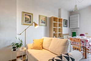 Una sala de estar con un sofá, una mesita con una lámpara, una estantería y un comedor con una mesa y sillas.