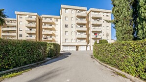 Um edifício residencial de vários andares com entrada central, rodeado de vegetação.