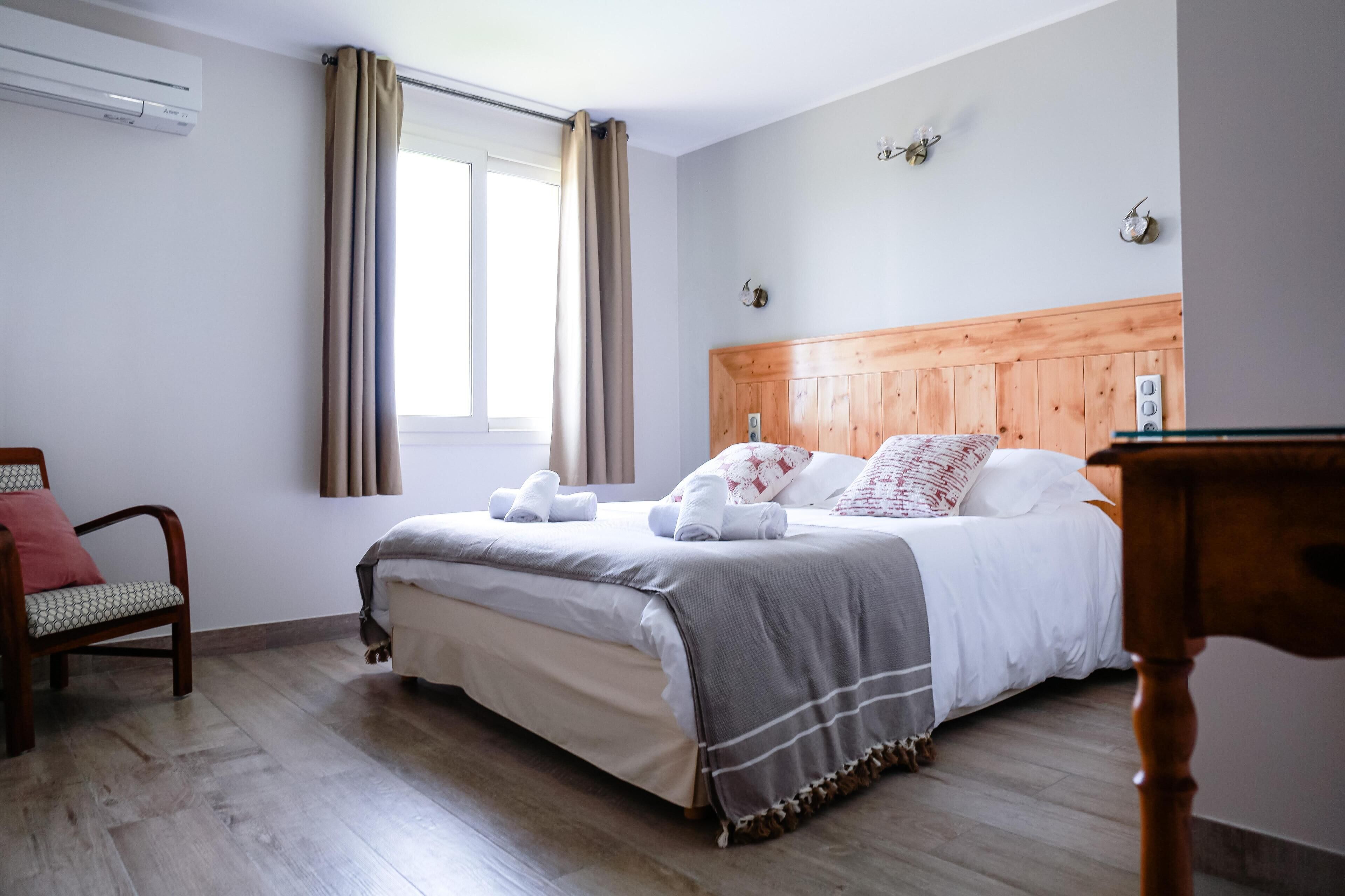 A bedroom with a wooden headboard, a bed with white bedding and a grey blanket, a wooden chair, and a window with curtains.