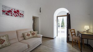 Een woonkamer met een bank, een bureau en een stoel. Er hangt een schilderij aan de muur en een deur die naar buiten leidt.