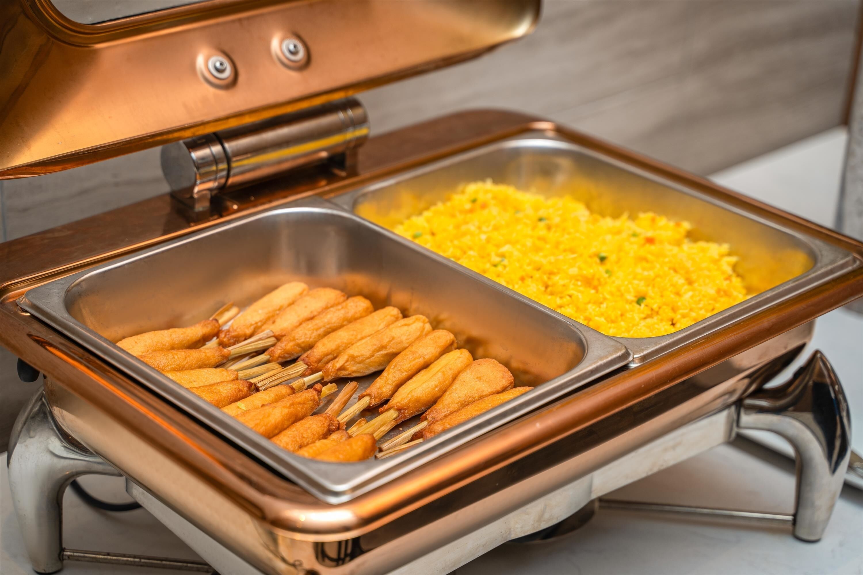 A buffet with two metal serving trays, one containing corn dogs and the other with yellow rice.