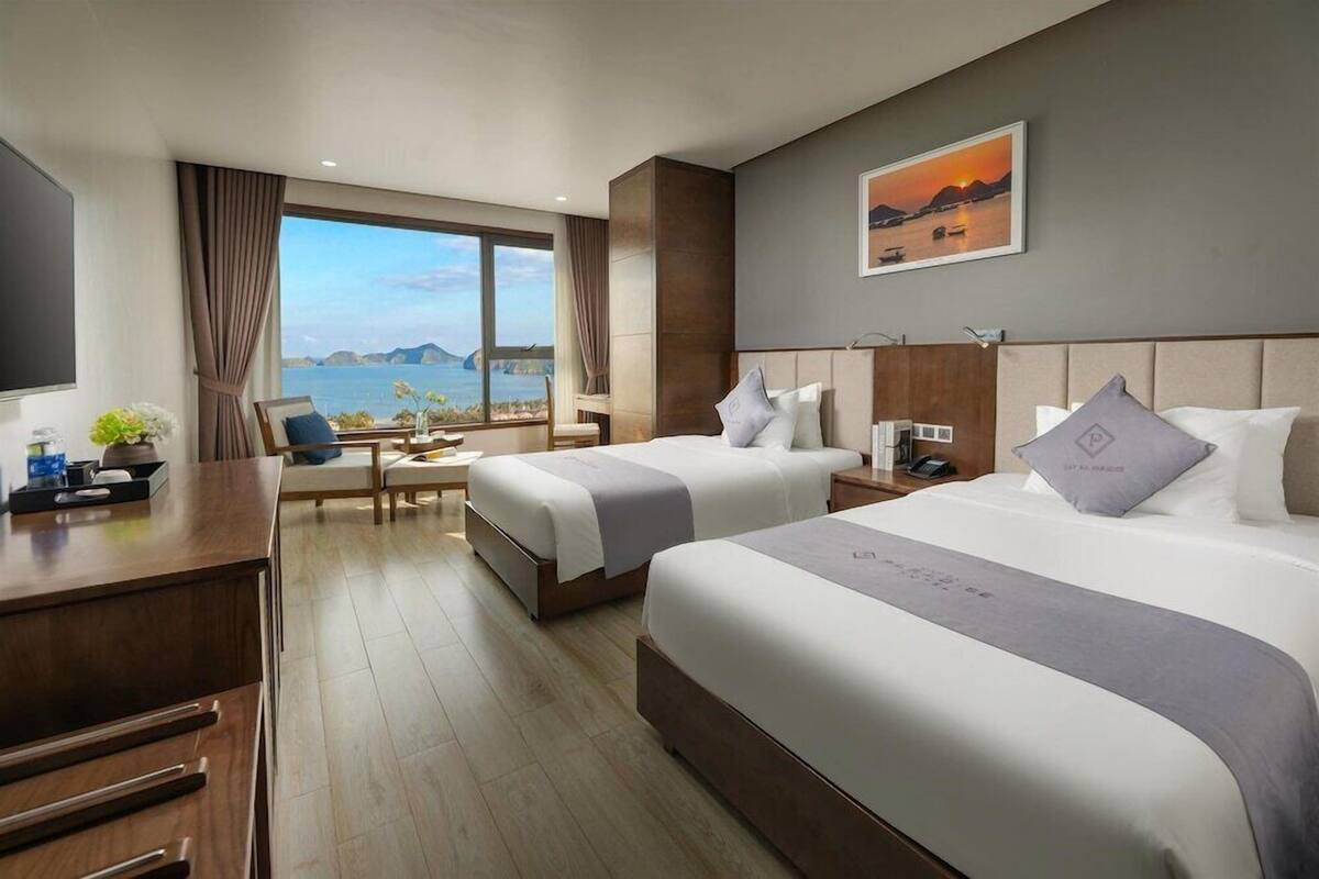A hotel room with two beds, a desk, a chair, a TV, and a large window with a view of the sea.