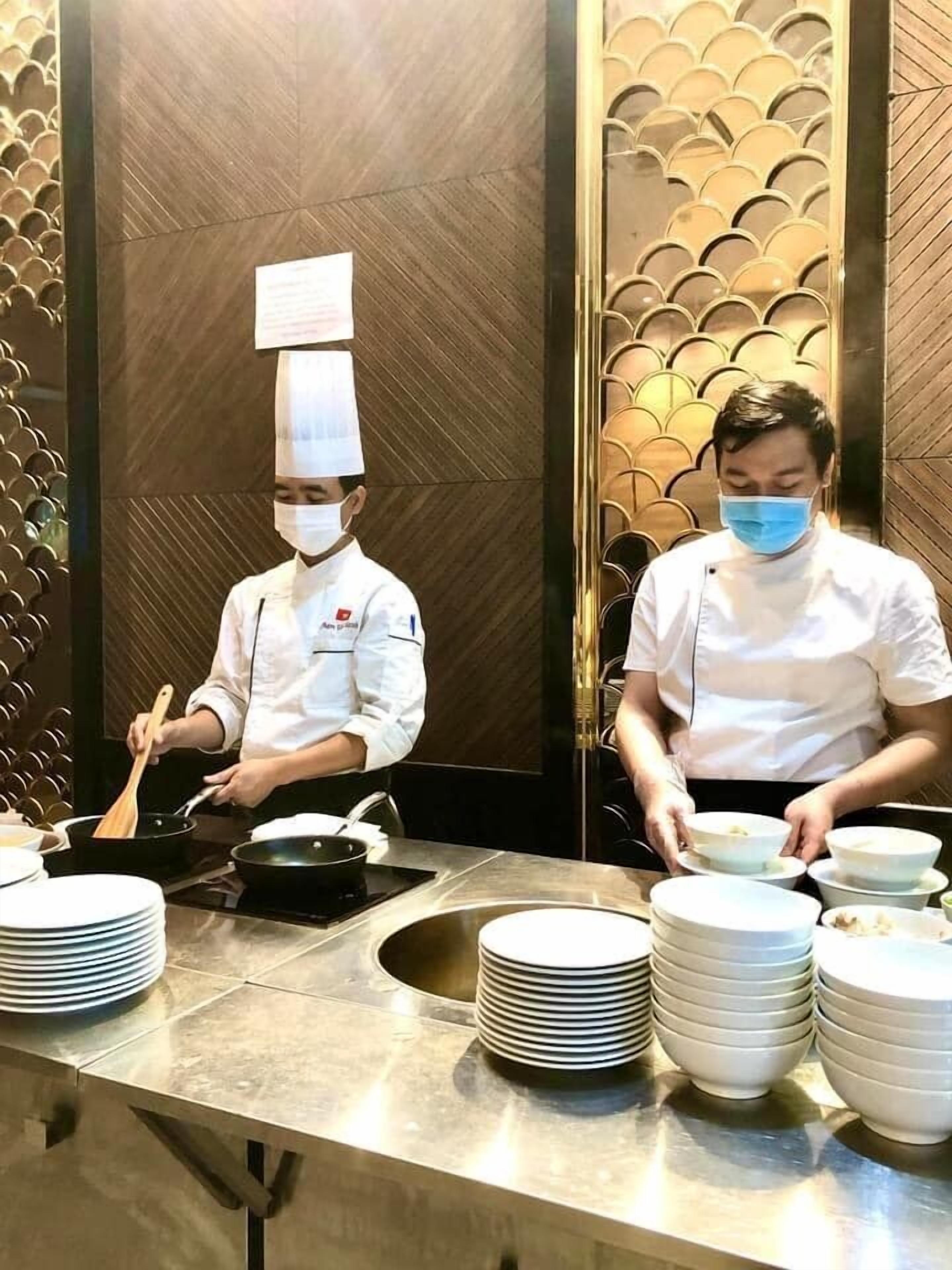 A professional kitchen with chefs preparing food, stainless steel tables, and a decorative wall with a fish scale pattern.