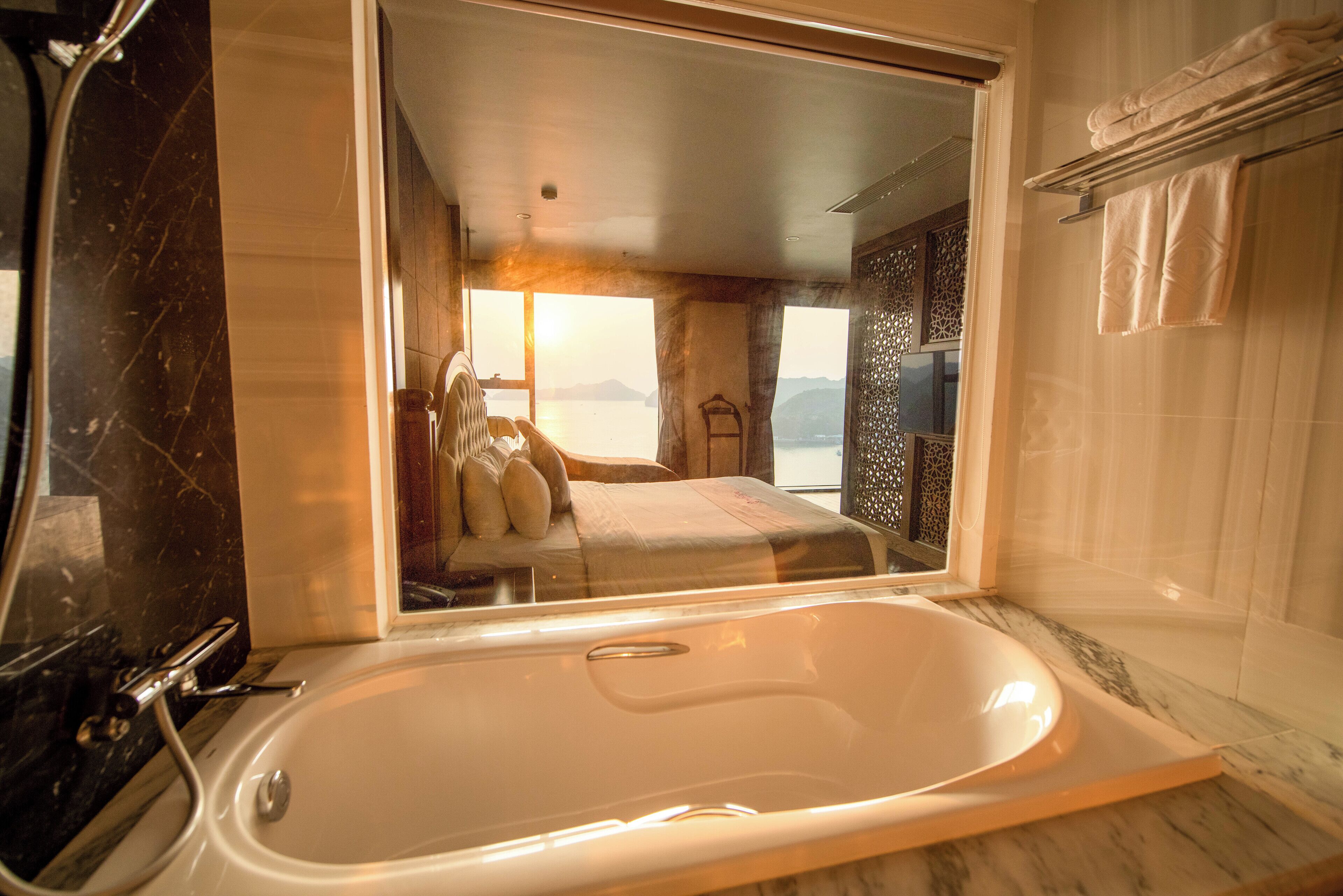 Un baño moderno con una bañera grande, zona de ducha y vista a un dormitorio con cama y sillón.