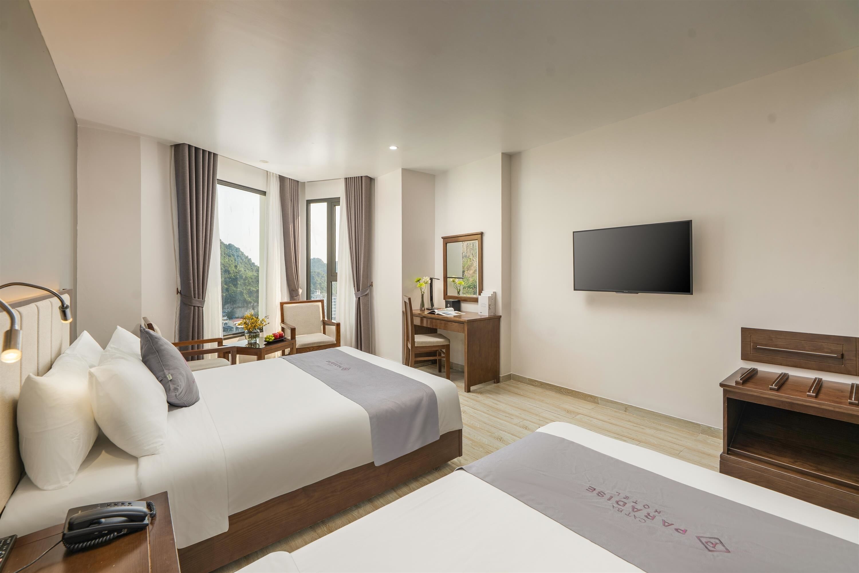 Habitación de hotel con cama, mesita de noche, lámpara, escritorio, silla, televisor y vistas al exterior con vegetación.