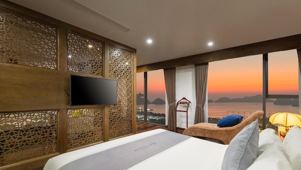 A hotel room with a bed, a TV, a view of the sunset, and a balcony chair. - Cat Ba Paradise Hotel - Sky Bar & Massage  (Hai Phong)