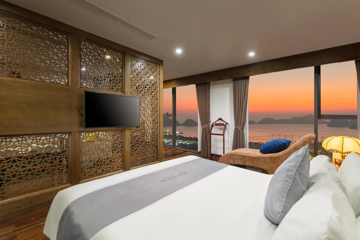 A hotel room with a bed, a TV, a view of the sunset, and a balcony chair.
