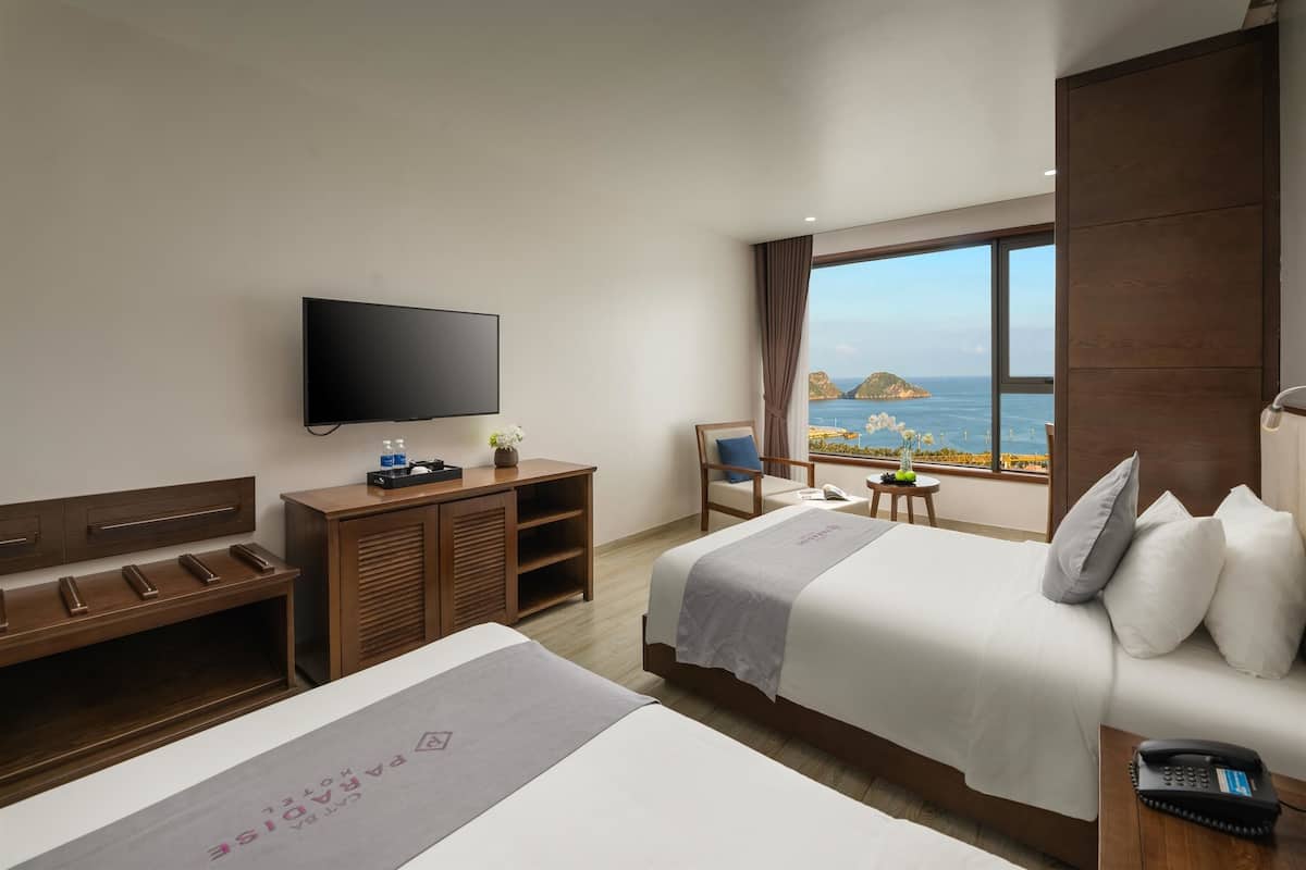 A hotel room with a TV, two beds, a desk, a chair, and a view of the sea.