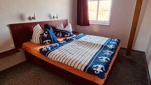 Ein ordentlich bezogenes Bett mit gestreifter Decke und Kissen, ein hölzernes Kopfteil, eine kleine Ablage und ein Fenster mit Vorhängen.