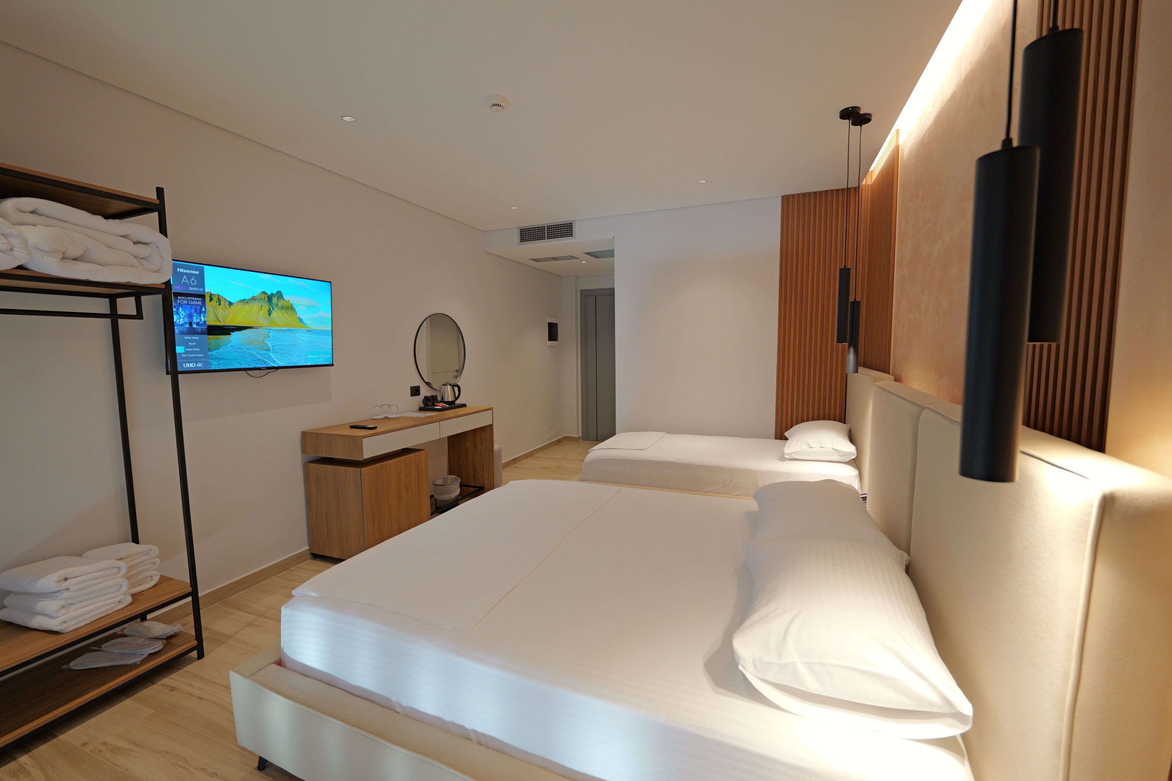 A modern hotel room with a large bed, a TV, a wooden shelf with towels, a desk, and a mirror.